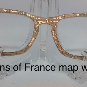 May include: Gold-toned glasses with a map of France printed on the frame. The map is in shades of beige and green with gold foil accents. "Regions of France map with foil" is printed below the glasses.