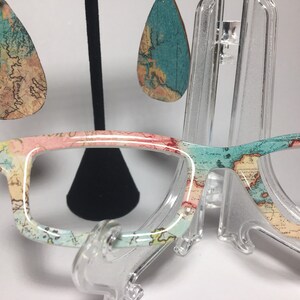 May include: A pair of eyeglasses with a map print design. The frames are white with a pink and blue map pattern. The eyeglasses are on a clear plastic stand. There are also a pair of teardrop-shaped earrings with a map print design.