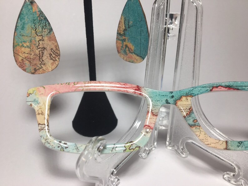 May include: A pair of eyeglasses with a map print design. The frames are white with a pink and blue map pattern. The eyeglasses are on a clear plastic stand. There are also a pair of teardrop-shaped earrings with a map print design.