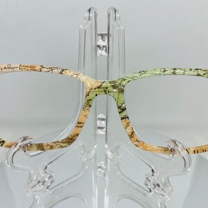 May include: A pair of eyeglasses with a vintage map print design. The frames are a light brown color with a green and beige pattern. The lenses are clear.