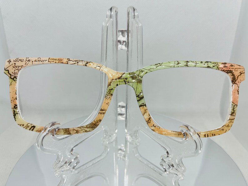 May include: A pair of eyeglasses with a vintage map print design. The frames are a light brown color with a green and beige pattern. The lenses are clear.