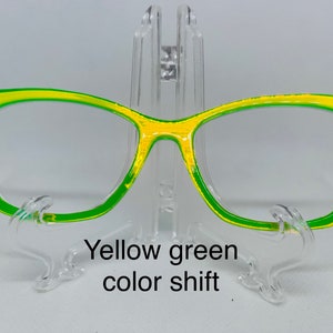 May include: A pair of cat-eye glasses with a yellow and green color shift. The glasses are on a clear plastic stand.