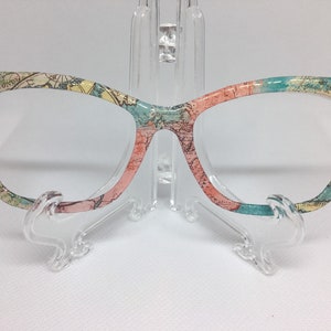 May include: Cat-eye glasses with a world map print in shades of blue, green, and pink. The glasses are on a clear plastic stand.