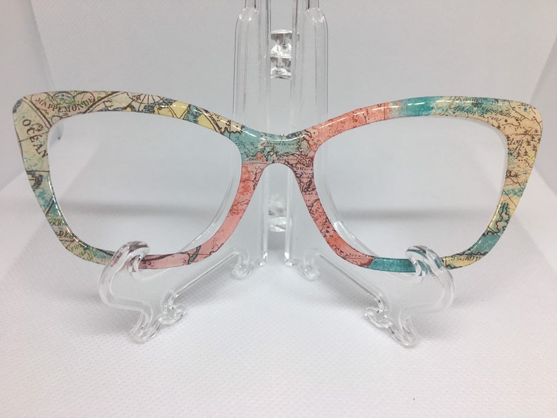 May include: Cat-eye glasses with a world map print in shades of blue, green, and pink. The glasses are on a clear plastic stand.