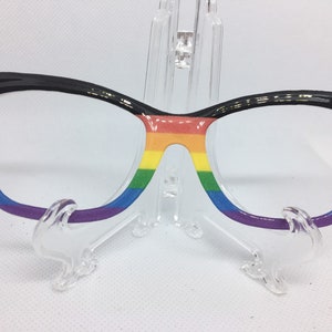 May include: Black cat-eye glasses with a rainbow design on the frames. The rainbow colors are red, orange, yellow, green, blue, indigo, and violet.