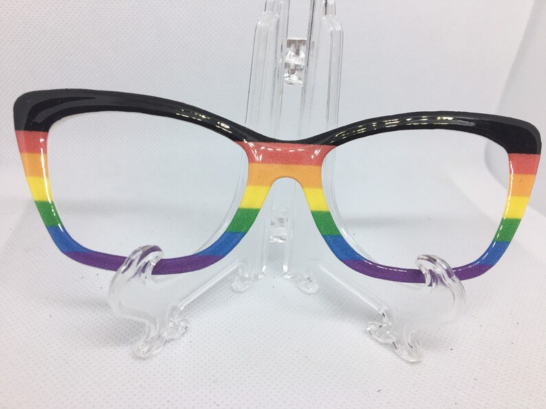 May include: Black cat-eye glasses with a rainbow design on the frames. The rainbow colors are red, orange, yellow, green, blue, indigo, and violet.