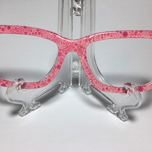 May include: Pink eyeglasses with a pink and white pattern of hearts, stars, and ribbons. The glasses are on a clear plastic stand.