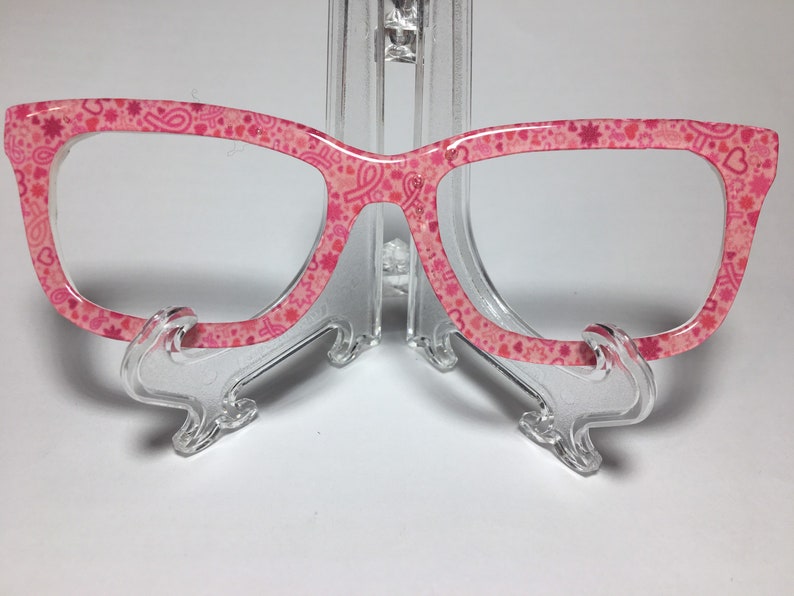 May include: Pink eyeglasses with a pink and white pattern of hearts, stars, and ribbons. The glasses are on a clear plastic stand.