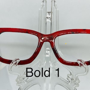 May include: Red square-shaped eyeglasses with a bold, marbled pattern. The glasses are on a clear plastic stand. The text "Bold 1" is visible below the glasses.