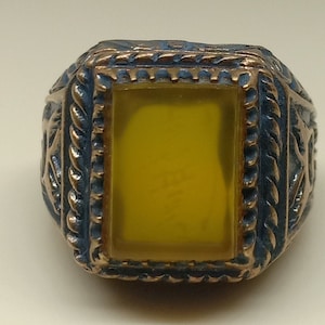 May include: A gold ring with a yellow stone set in a square bezel. The ring has an intricate design around the stone.