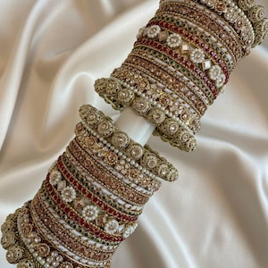 Peut inclure: Un ensemble de deux piles de bracelets rigides dorés ornés de travail du métal complexe, de petites perles blanches et de pierres rouges et vertes. Les bracelets présentent des textures variées, des bords perlés et des motifs floraux, avec bokoraJewels.com.