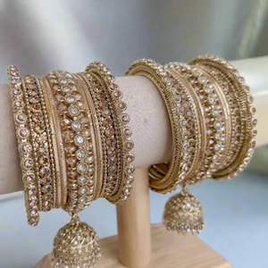 May include: Gold-toned bangles with intricate designs and embedded crystals. The bangles are displayed on a wooden stand. Each set includes multiple bangles, some with dangling bell-shaped charms. The jewelry has a warm, radiant appearance.