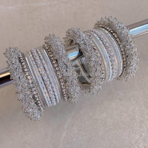 May include: A collection of white and silver bangles, adorned with detailed beadwork and sparkling embellishments. The bangles are displayed on a silver rod, highlighting diverse textures and designs, including pearl-like beads and shimmering elements. The jewellery is elegant and visually appealing.