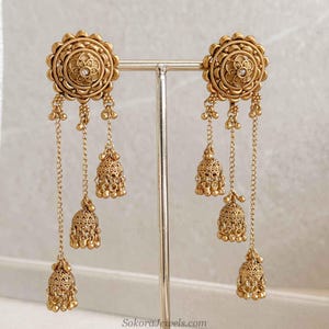 May include: A pair of gold-toned earrings featuring a circular stud with an intricate floral pattern and a central clear stone. Three delicate chains of varying lengths hang from each stud, each ending in a small, ornate bell-shaped charm.