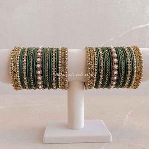 May include: A set of dark green bangles featuring gold-tone metal accents, small white faux pearls, and clear crystals. The bangles are arranged in two stacks on a display stand, with the text SokoraJewels.com visible between the two stacks.