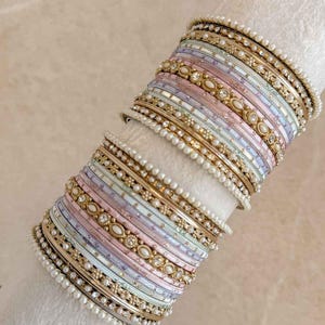 May include: A collection of pastel-coloured bangles with gold detailing and faux pearls. The bangles feature pink, blue, and lavender hues, with decorative gold bands and pearl embellishments. The jewellery is presented on a white surface.