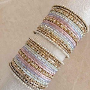 May include: A stack of bangles featuring a mix of gold-tone metal, pastel pink, blue, and lavender enamel bands. The set includes thin bands adorned with small white faux pearls, textured gold-tone metal rings, and wider bands with embedded clear stones.