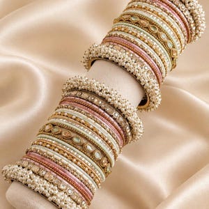 Könnte beinhalten: Ein Set aus gestapelten Armreifen mit abwechselnden Bändern in Rosa, Mintgrün und goldfarbenem Metall. Die Kollektion umfasst strukturierte Metallringe, Reihen kleiner weißer Kunstperlen und eingelassene klare Steine in einem Muster.