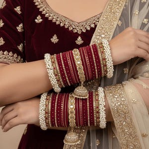 Pode incluir: Um conjunto de pulseiras de veludo bordô com detalhes em metal dourado, missangas intrincadas e pequenos pingentes em forma de sino. As pulseiras são bordadas com contas brancas tipo pérola e usadas com uma blusa de veludo bordô a combinar.
