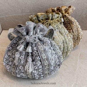 Peut inclure: Trois pochettes à cordon ornées de broderies complexes, de fils métalliques argentés et dorés, et de perles denses. Les sacs sont disposés en rangée, présentant une pochette argentée devant, suivie d'une dorée et d'une autre couleur bronze.
