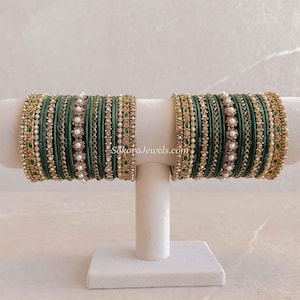 Puede incluir: Un conjunto de brazaletes de color verde oscuro y dorado dispuestos en un soporte de exhibición. Los brazaletes presentan intrincados trabajos en metal dorado, pequeñas perlas blancas y detalles verdes. El texto SokoraJewels.com es visible.