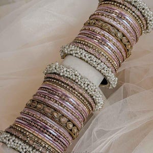 May include: A set of stacked bangles in shades of light pink, gold, and white. The collection features metallic gold bands, smooth pink enamel rings, and decorative bands encrusted with small white pearls and pink teardrop-shaped stones.