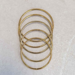 May include: Several gold-toned bangles, each featuring a line of tiny, closely-set beads. The bangles are arranged in a layered formation, highlighting their circular shape. The backdrop is a plain, light-coloured surface. The website address SokoraJewels.com is visible.