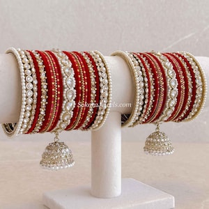 Op de afbeelding: Een set van twee bijpassende armbanden in dieprood fluweel, voorzien van goudkleurige metalen accenten, rijen witte kunstparels en heldere steentjes. Elke set bevat een bungelende goudkleurige jhumka-bedel met witte kraaldetails.