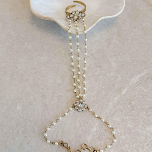 Op de afbeelding: Een goudkleurige handketting bestaande uit een ring die met drie fijne strengen van kleine witte parels verbonden is aan een armband. De ring en het centrale verbindingsstuk zijn versierd met heldere, gefacetteerde stenen. De tekst SokoraJewels.com is zichtbaar.