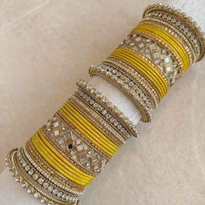 May include: A set of yellow and gold bangles featuring intricate metallic textures, small clear rhinestones, and diamond-shaped mirror accents. The stack includes multiple thin yellow thread-wrapped bands and decorative beaded edges. Text reads SokoraJewels.com.