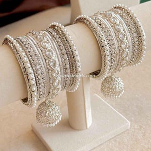 May include: A set of silver-toned bangles featuring white enamel, clear crystals, and small white pearls. The bracelets include intricate metalwork and a dangling bell-shaped charm with the text SokoraJewels.com visible on the central band.