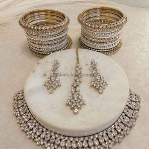 May include: A glamorous jewellery set comprising a necklace, earrings, a headpiece, and bangles. The set includes a sparkling necklace, matching earrings, and a headpiece, all embellished with clear gemstones. Two stacks of gold and white bangles complete the ensemble.
