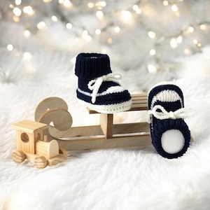 Op de afbeelding: Een paar marineblauwe en witte gebreide babyschoentjes met witte veters. De schoentjes staan op een kleine houten slee, naast een houten speelgoedtrein. De achtergrond is zacht, wit en wazig.