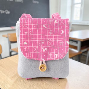 Könnte beinhalten: Ein grauer Filzrucksack mit einer rosa Klappe mit weißem Gittermuster und verschiedenen Symbolen. Der Rucksack hat rosa Träger und einen Holzknopfverschluss. Die Tasche steht auf einem Holztisch.