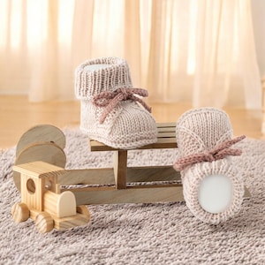 Op de afbeelding: Een paar gebreide babyschoentjes in een lichtroze tint, met een bijpassende strik. De schoentjes worden getoond op een kleine houten slee, naast een houten speelgoedtrein. De achtergrond heeft een zacht, neutraal getint tapijt en gordijnen.