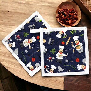 Könnte beinhalten: Zwei marineblaue, gesteppte Topflappen mit weißem Rand. Der Stoff zeigt ein sich wiederholendes Muster von Cartoon-Köchen, Küchenutensilien und Lebensmitteln. Der Text "Yes Chef!" und "Gourmet Delight" sind ebenfalls sichtbar.