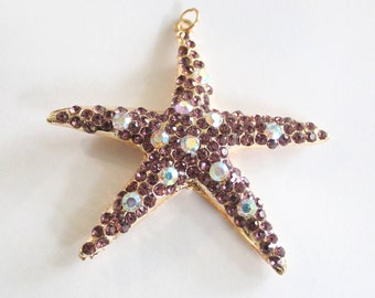 May include: A gold-coloured starfish pendant, adorned with small, purple rhinestones and larger, iridescent crystals. The pendant has a loop at the top for hanging. The starfish design is a popular theme for jewellery and accessories.