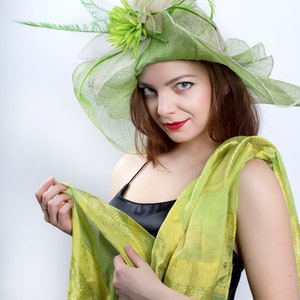 May include: A woman wearing a green and white hat with a flower and a green scarf. She is looking at the camera with a slight smile.