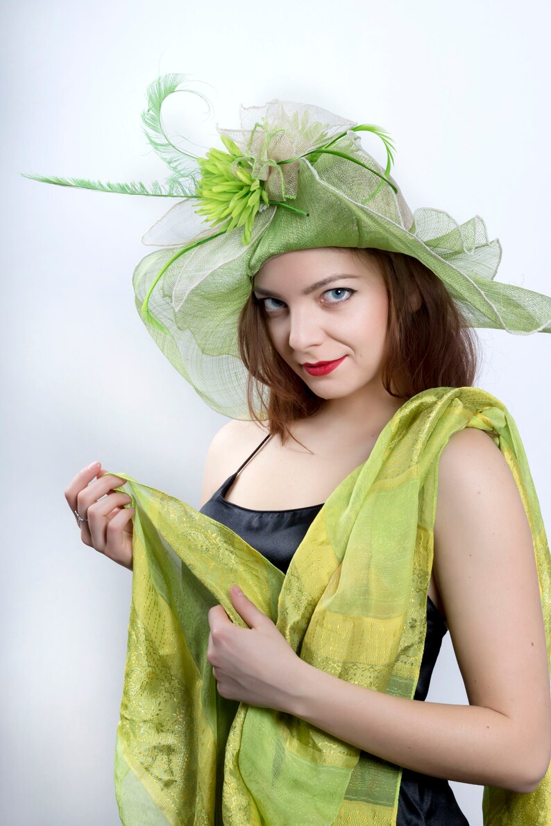 May include: A woman wearing a green and white hat with a flower and a green scarf. She is looking at the camera with a slight smile.