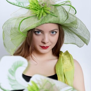 May include: A woman wearing a green and white hat with a green flower and a green scarf. The hat is made of a mesh material and has a wide brim.