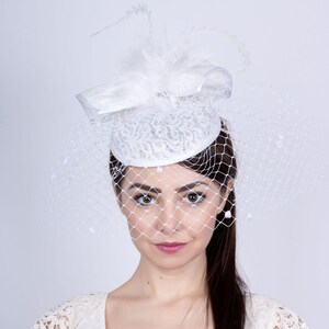 May include: A white bridal hat with a veil and a small white feather accent. The hat is made of lace and has a round shape.