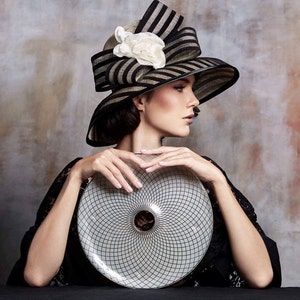 May include: A woman wearing a black and white striped hat with a white flower accent holds a white and black circular plate with a geometric design.