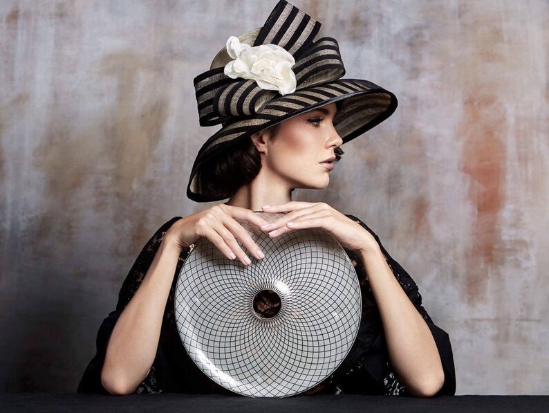 May include: A woman wearing a black and white striped hat with a white flower accent holds a white and black circular plate with a geometric design.