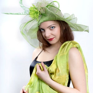 May include: A woman wearing a green and white hat with a flower and a green and yellow scarf. She is wearing a black sleeveless dress.