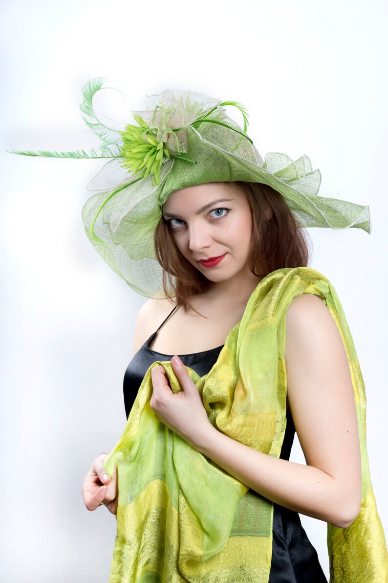May include: A woman wearing a green and white hat with a flower and a green and yellow scarf. She is wearing a black sleeveless dress.