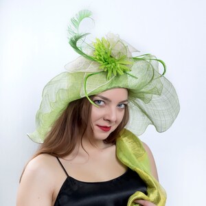 May include: A woman wearing a green and white hat with a flower and feather embellishment. The hat is made of a delicate, sheer fabric. She is also wearing a black top and a green scarf.