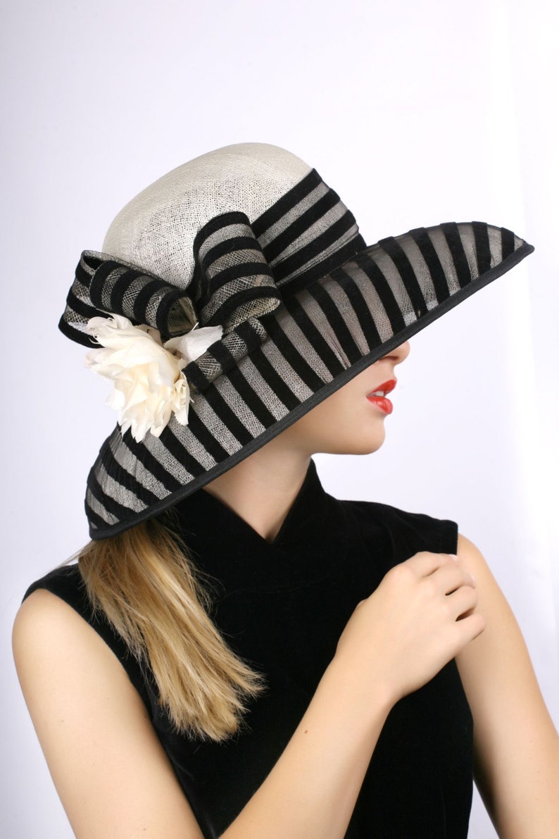 May include: A black and white striped hat with a large white flower accent. The hat is worn by a person with blonde hair and red lipstick. The person is wearing a black sleeveless dress.