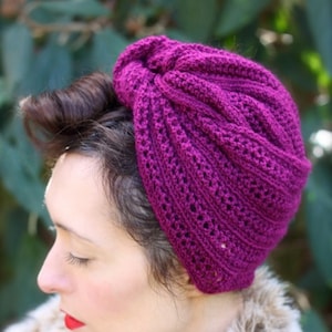 May include: A close-up of a woman wearing a deep purple knitted turban-style hat. The hat has a loose, open weave and is styled in a way that creates a dramatic, voluminous look.