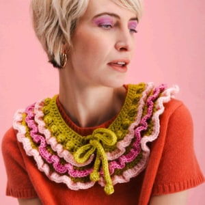 Puede incluir: Una mujer lleva un jersey naranja brillante con un cuello de ganchillo. El cuello está hecho de tres capas de volantes en hilo rosa, verde y amarillo. El cuello tiene una corbata de lazo en la parte delantera.
