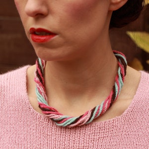 May include: A pink sweater with a multi-colored braided necklace. The necklace is made of brown, pink, and light blue yarn.